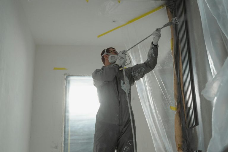 A professional painter wearing protective gear spray paints a wall indoors, working on a home renovation project.