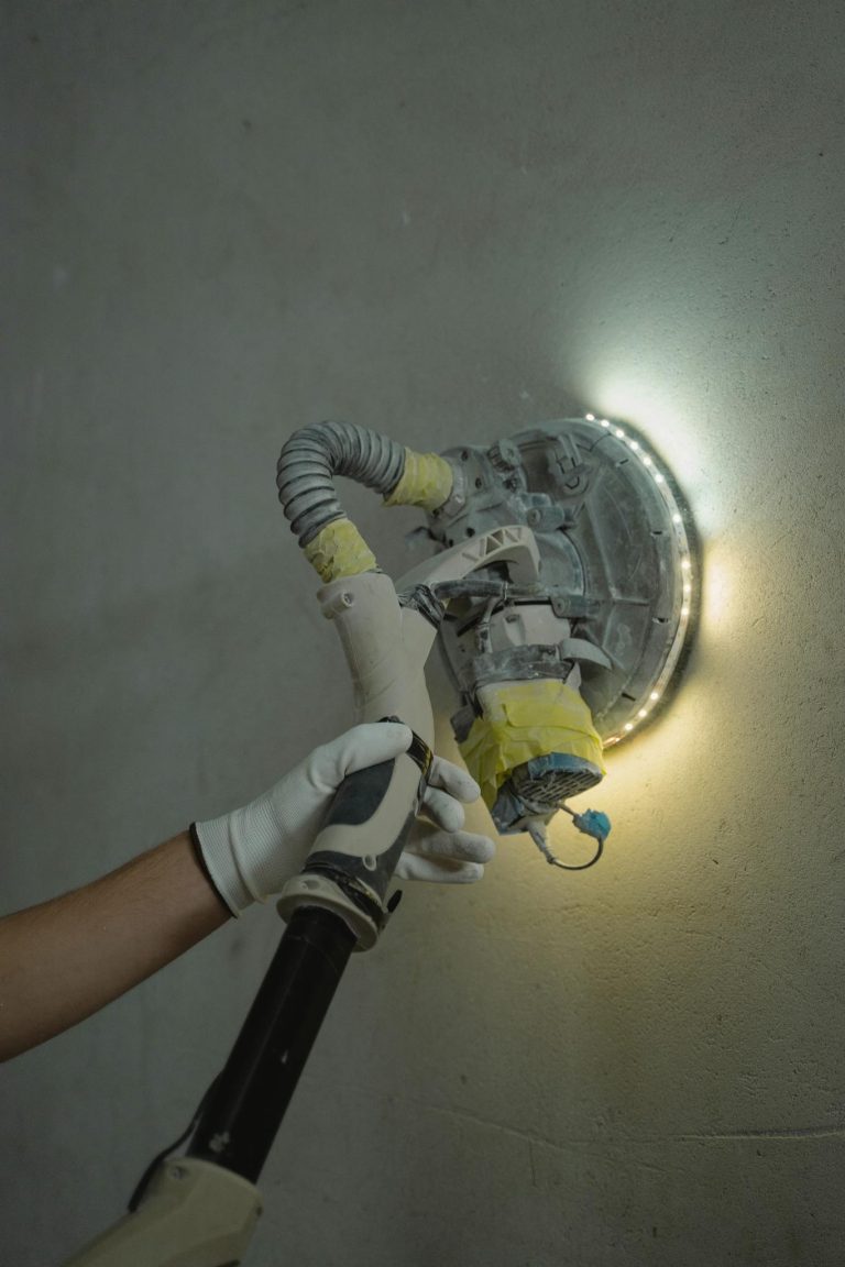 Close-up of a worker sanding a wall with a power tool under bright lighting.