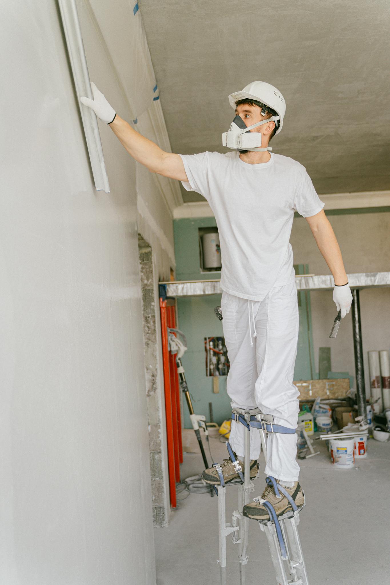 Construction worker on stilts painting a wall indoors, wearing protective gear and a hard hat.